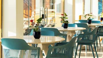 Een moderne eetruimte met ronde tafels en teal gekleurde stoelen, grote ramen met uitzicht op de stad en potplanten op de tafels.