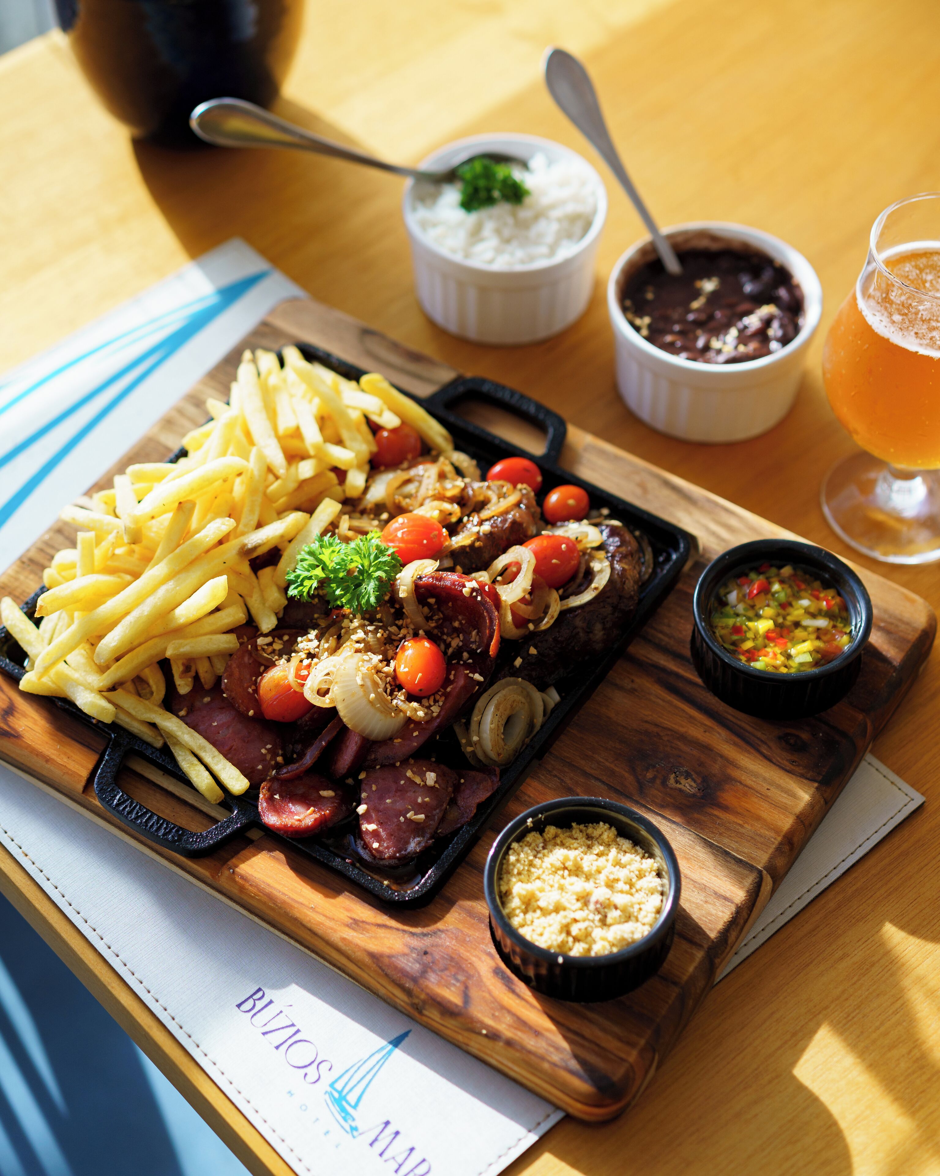 A wooden serving board with a steak, fries, and vegetables, accompanied by small bowls of condiments and a glass of beer.