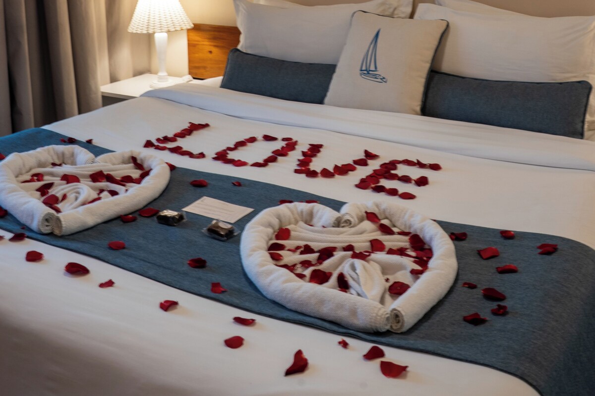A hotel room with a bed adorned with rose petals and towels spelling "LOVE".