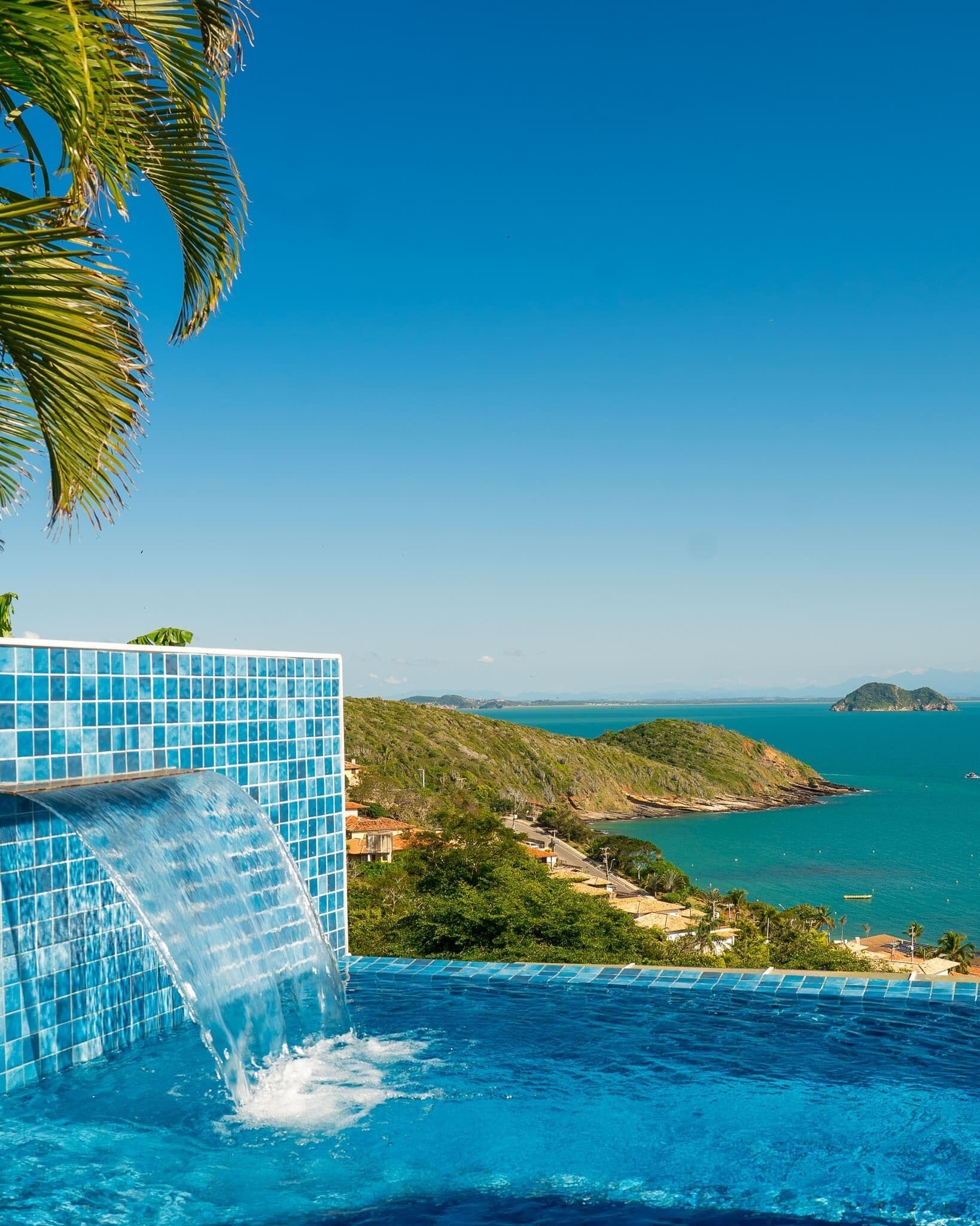 A pool with a waterfall feature, surrounded by a view of a coastal landscape with hills and a body of water.