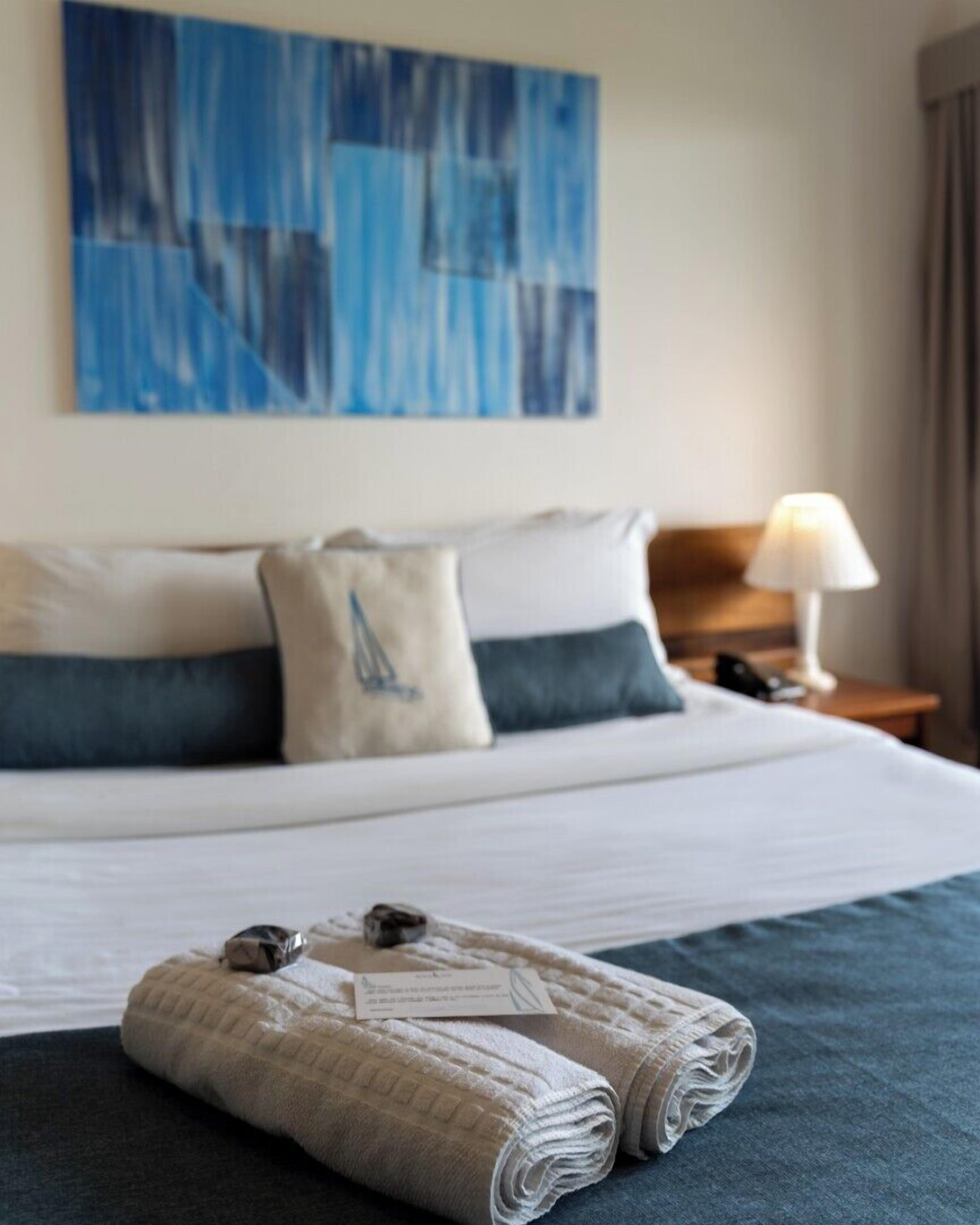 Una cama bien hecha con edredón azul y blanco, una lámpara en la mesita de noche y un cuadro abstracto azul en la pared.