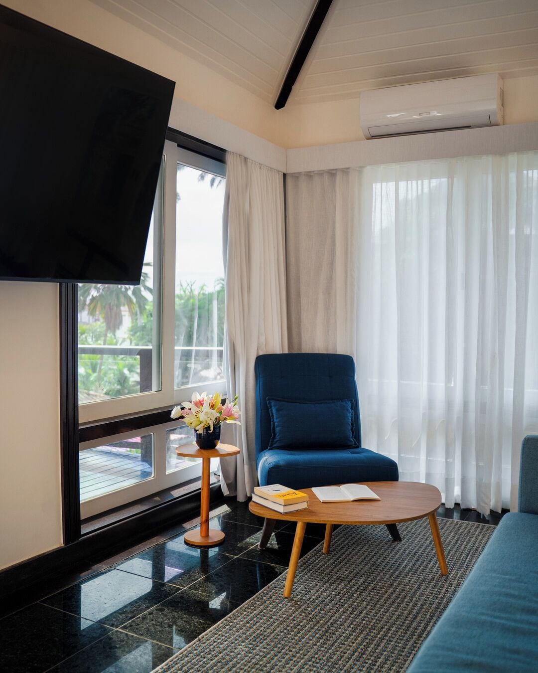 Habitació d'hotel moderna amb una finestra gran, un divà blau, una taula de fusta petita amb llibres i un vas de flors.
