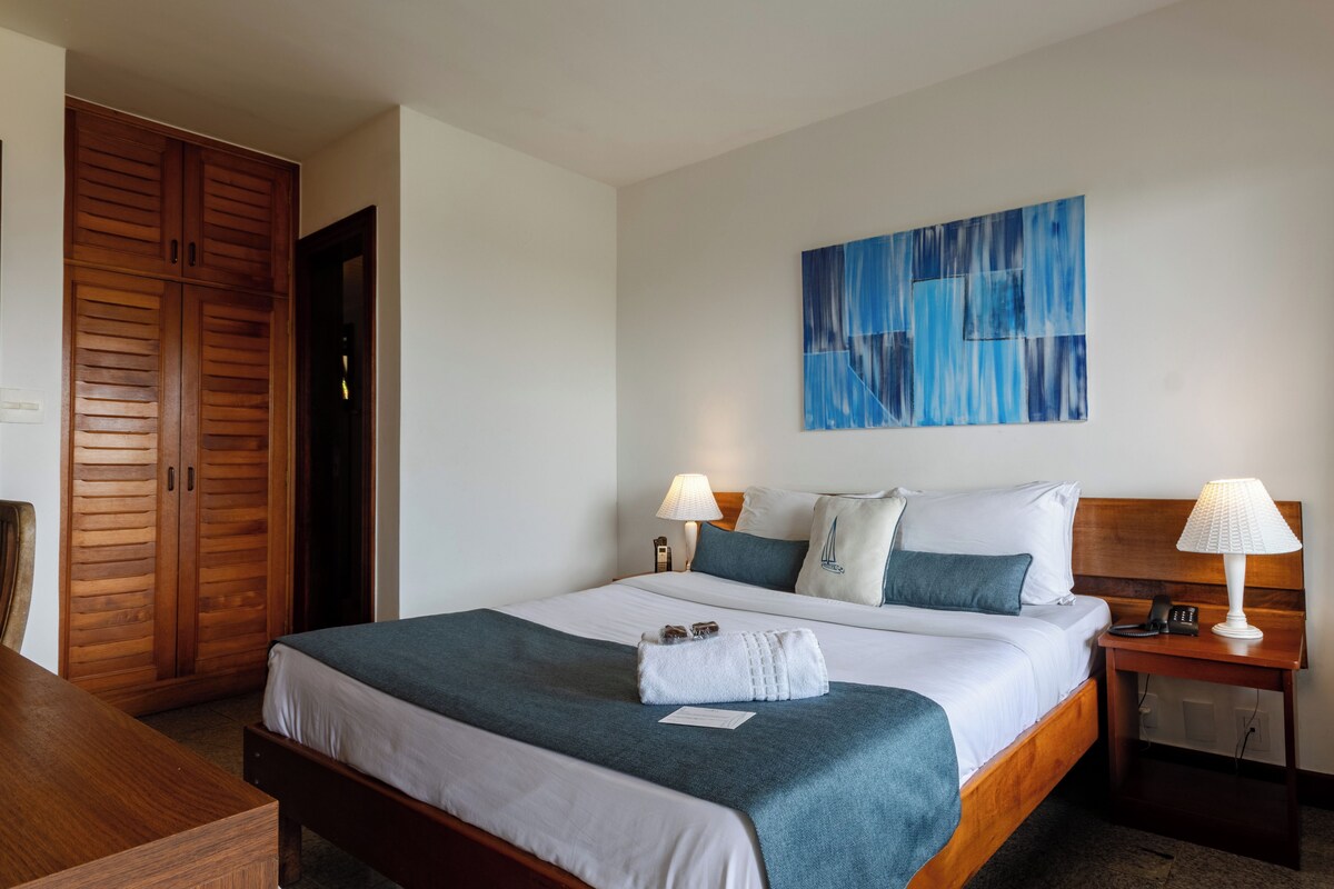 A hotel room with a bed, wooden wardrobe, bedside lamps, and a blue abstract painting on the wall.