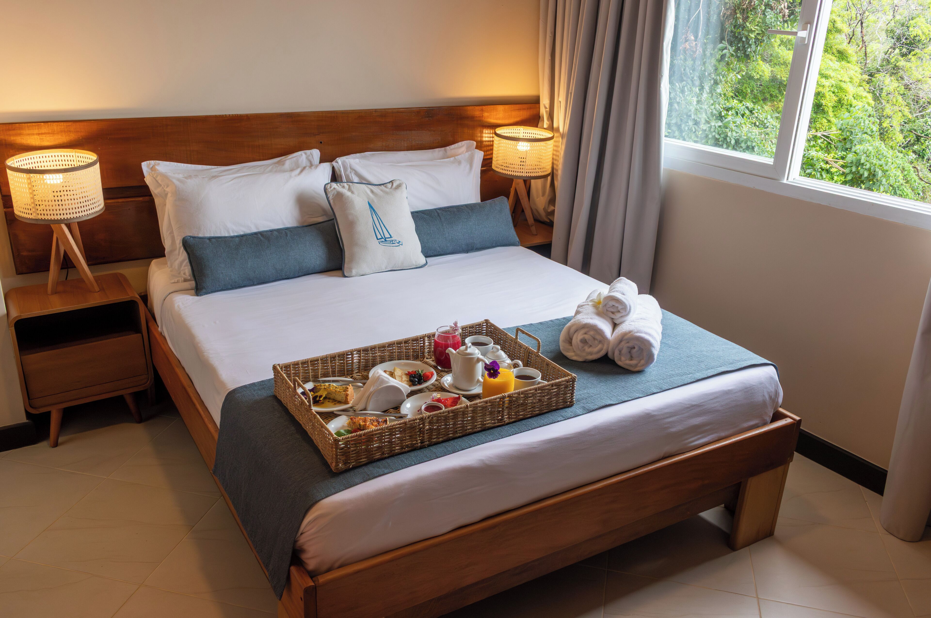 Un lit bien fait, avec un plateau contenant des articles pour le petit-déjeuner, une tête de lit en bois et une fenêtre donnant sur un paysage verdoyant.