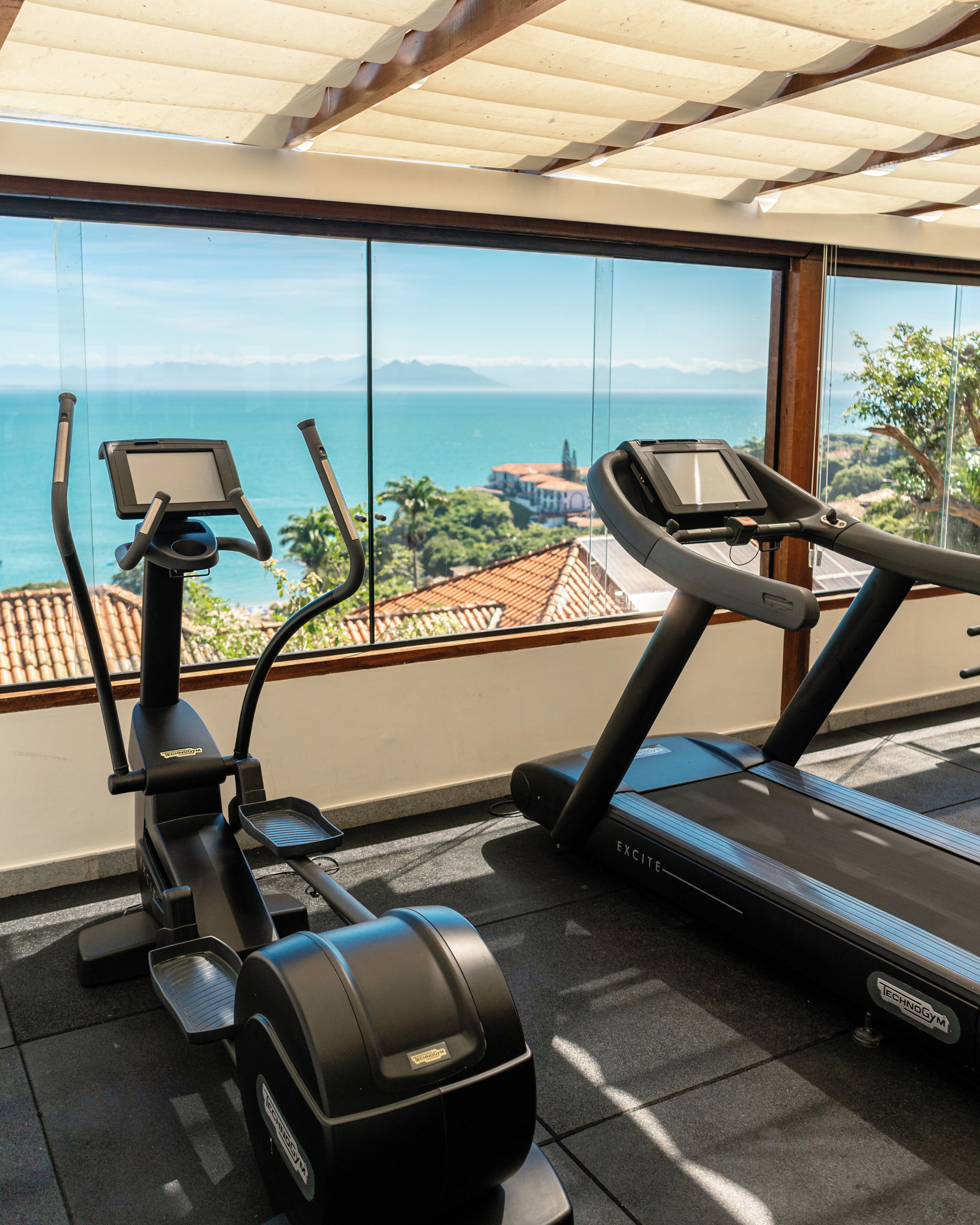 An indoor gym with exercise equipment, including a stationary bike and a treadmill, with a view of the sea and buildings through large windows.