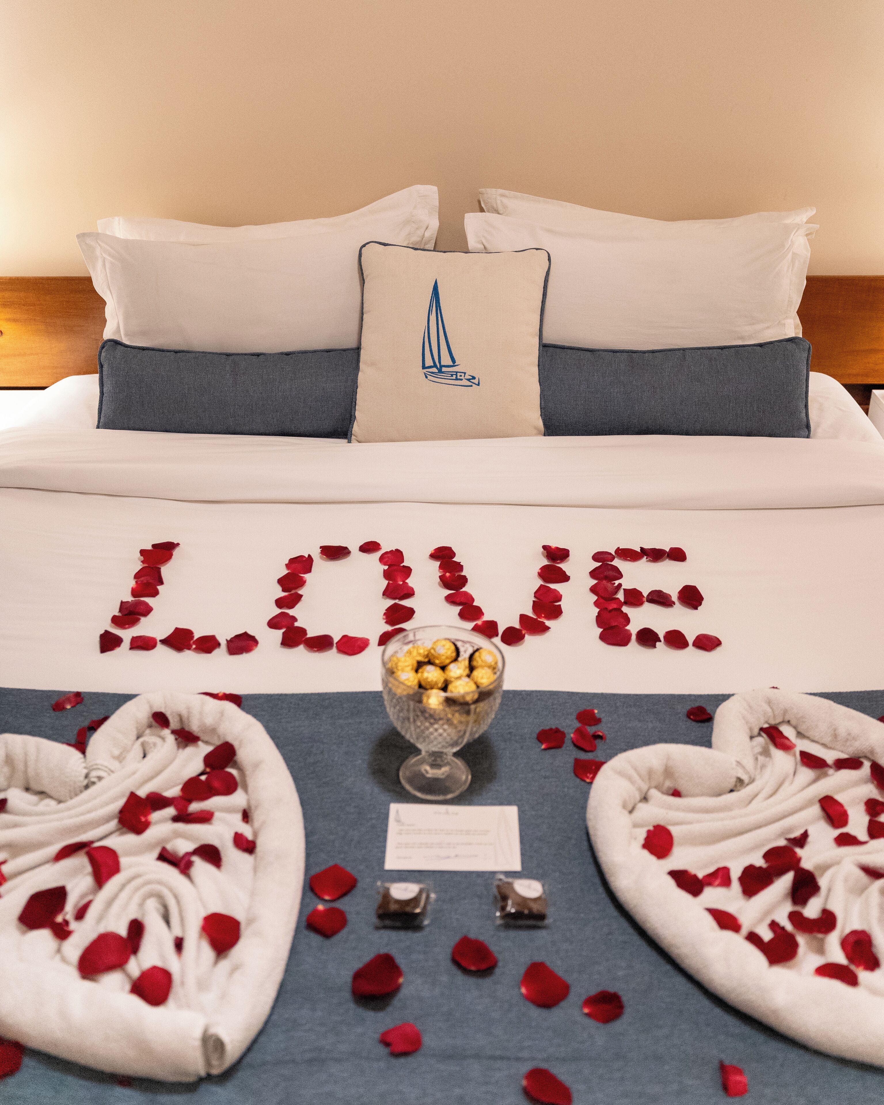 A bed with a 'LOVE' decoration made of rose petals, a glass bowl of chocolates, and a pillow with a sailboat design.