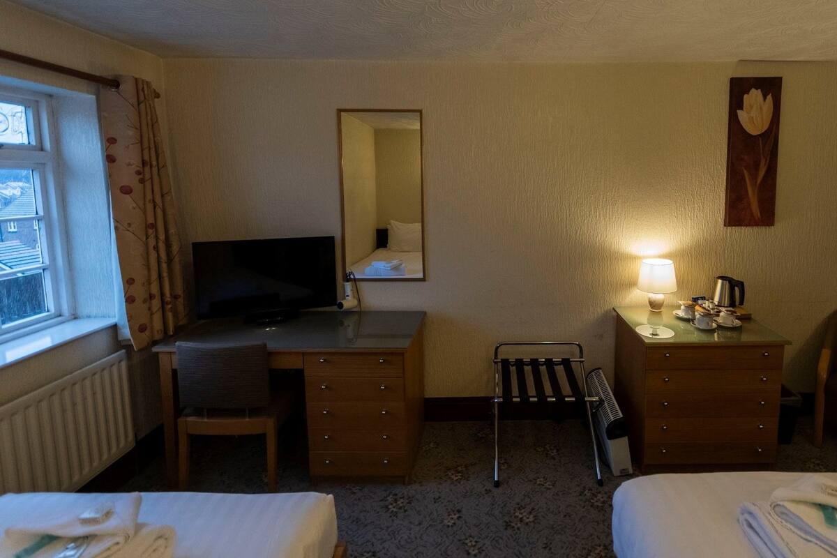 A hotel room with a bed, a desk with a TV, a chair, a small table with a lamp, and a radiator.