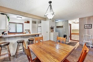A dining area with a wooden table and four chairs, a kitchen area with white cabinets and a refrigerator, and a hanging light fixture.