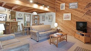 Un salon chaleureux avec un canapé, une table basse et un meuble pour la télévision. On y trouve également un coin repas avec des chaises et une table. Les murs et les poutres du plafond sont en bois.