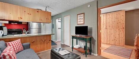 Un espacio habitable compacto con kitchenette, un sofá gris con almohadas rojas, un taburete negro, un televisor sobre un mueble de madera y un armario de madera.