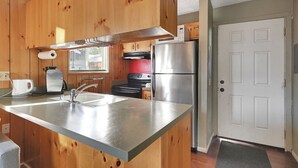 A compact kitchen with a stainless steel refrigerator, a built-in oven, and a microwave. There is a window with blinds, a sink with a faucet, and a coffee maker on the counter.