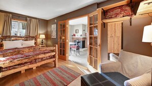A cozy room with a bed, a wooden bench, a small table with a lamp, and a view of a dining area through an open door.