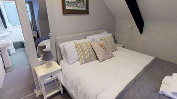 Un dormitorio con cama, mesitas de noche, lámpara, un cuadro en la pared y vista al baño.