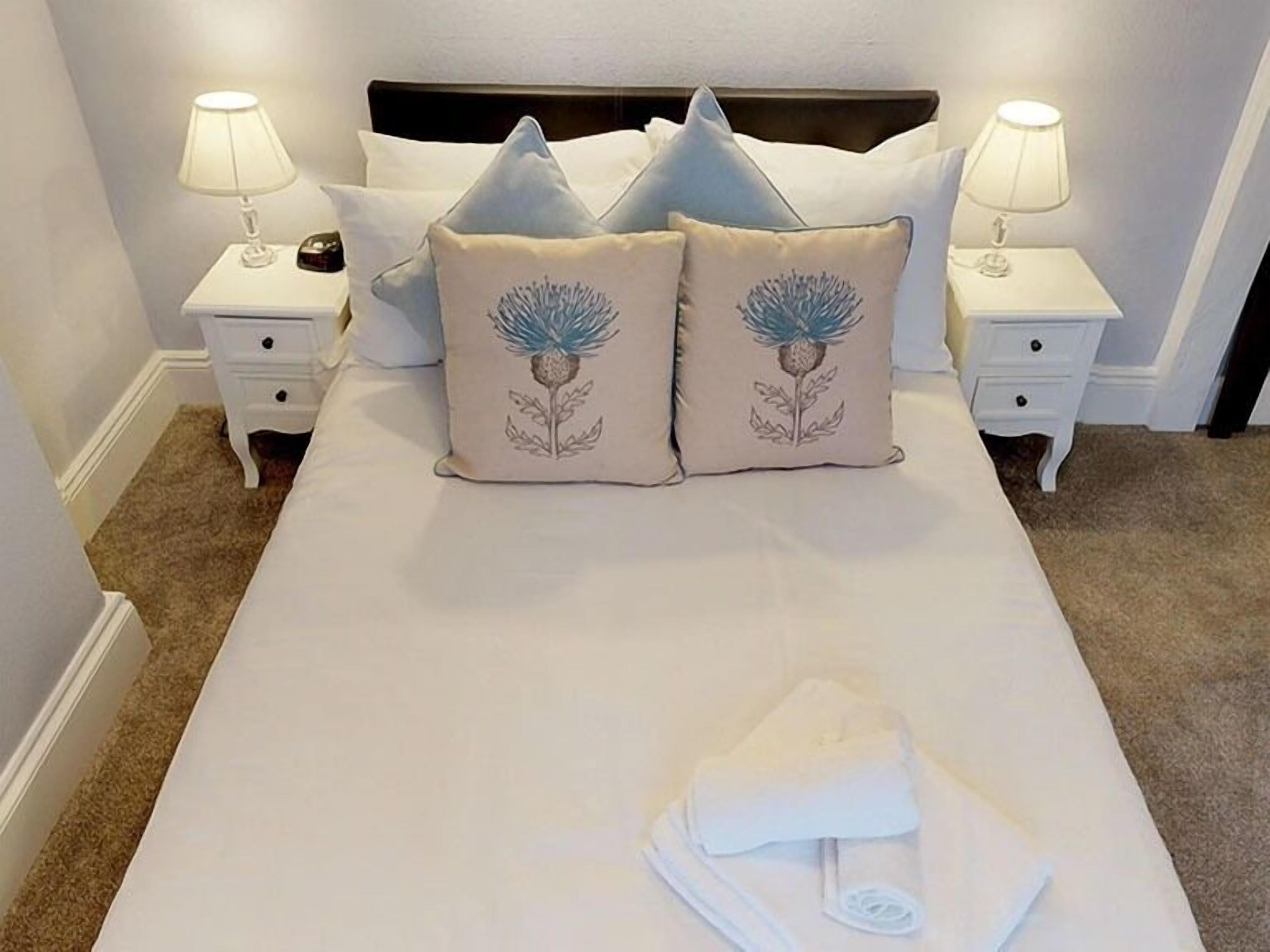 A neatly made bed with two decorative pillows, a white bedside table with a lamp, and a folded towel on the bed.