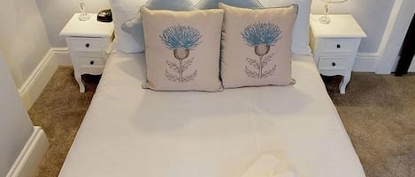 A neatly made bed with two decorative pillows, a white bedside table with a lamp, and a folded towel on the bed.