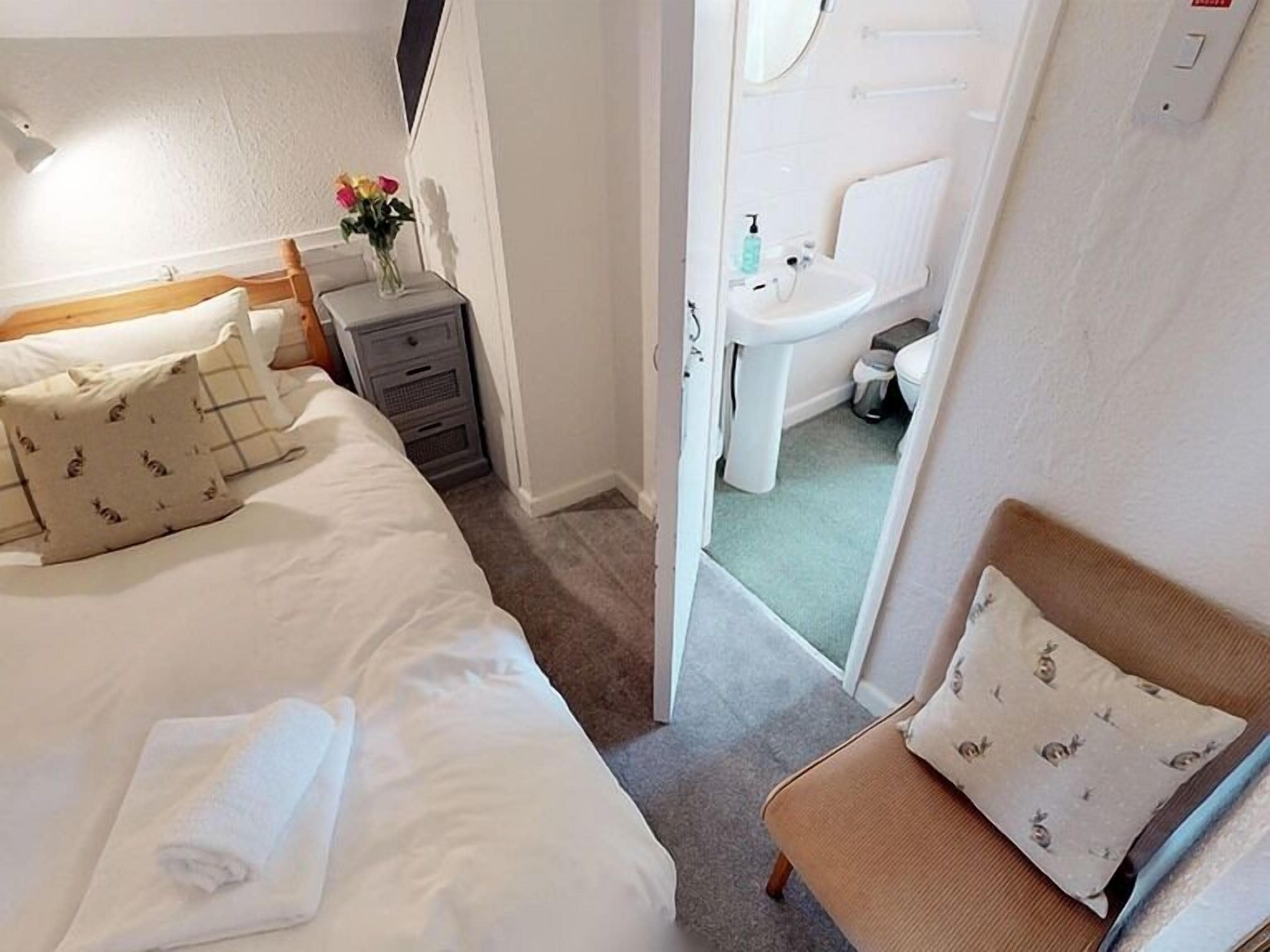 Un dormitorio con una cama, una mesita de noche con un florero, una silla y un baño visible a través de una puerta abierta.