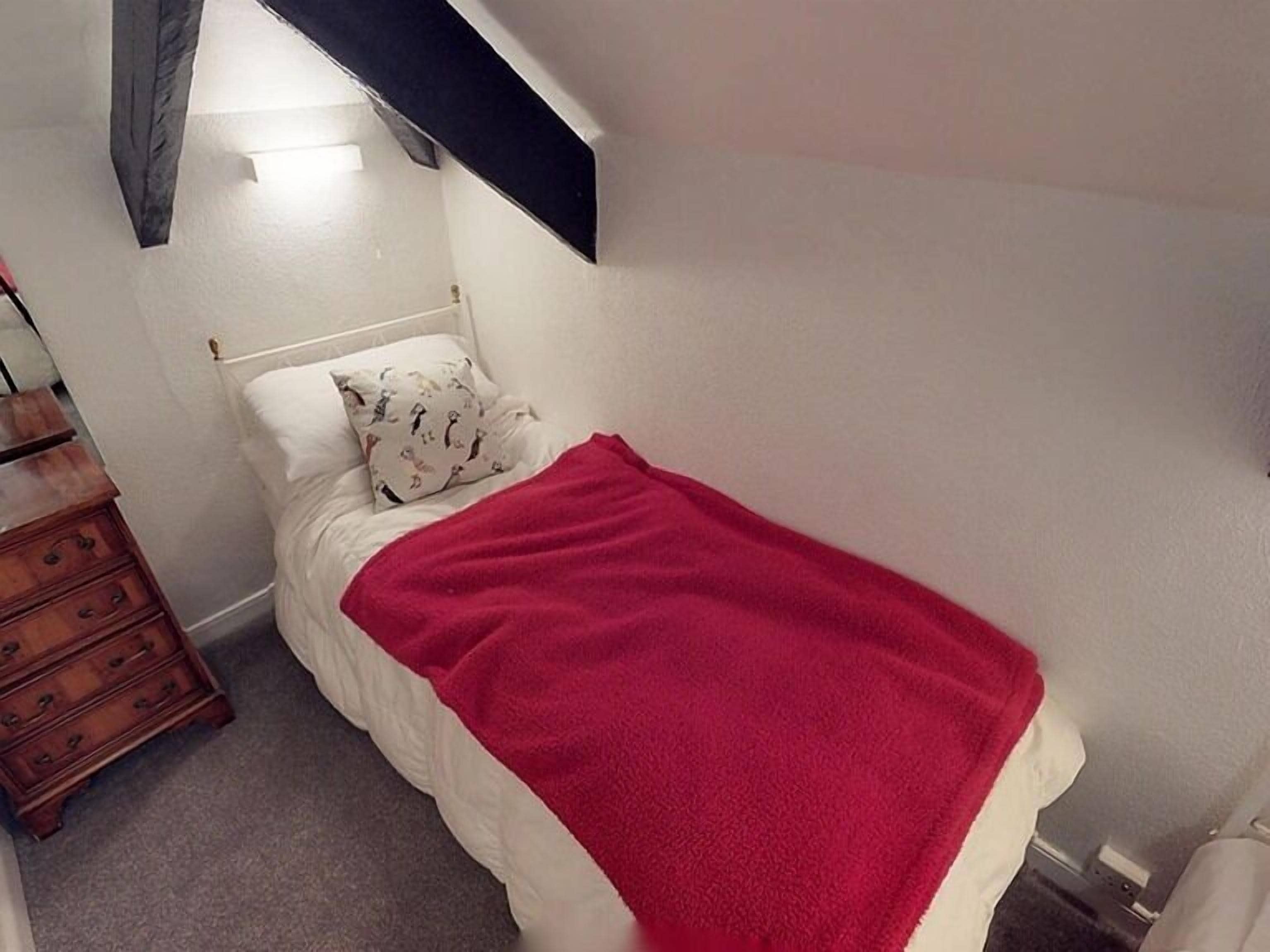 Un petit lit bien fait, avec une couverture rouge, une commode en bois et un oreiller blanc orné de motifs d’animaux.
