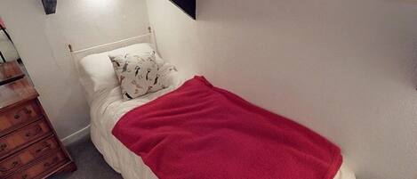 A small, neatly made bed with a red blanket, a wooden dresser, and a white pillow with animal prints.