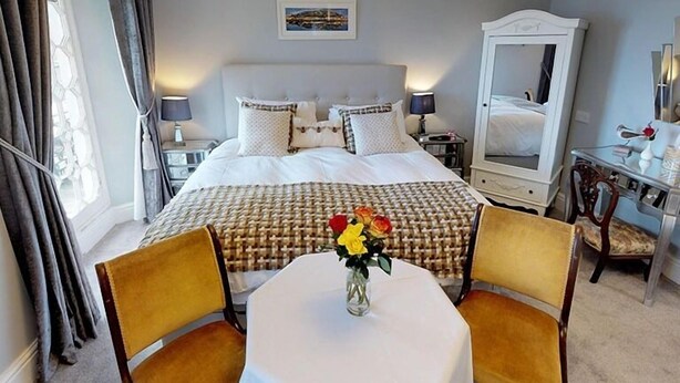 A hotel room with a large bed, a bedside table with a lamp, a desk with a chair, and a window with curtains. - Ranscombe House (Brixham)