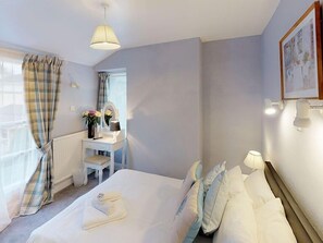 A hotel room with a bed, a bedside table with a lamp, a window with curtains, a radiator, and a framed picture on the wall.
