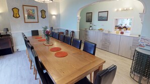 A dining area with a long wooden table, black chairs, and a vase of flowers. There is a kitchen area with a sink, stove, and cabinets. - Ranscombe House (Brixham)