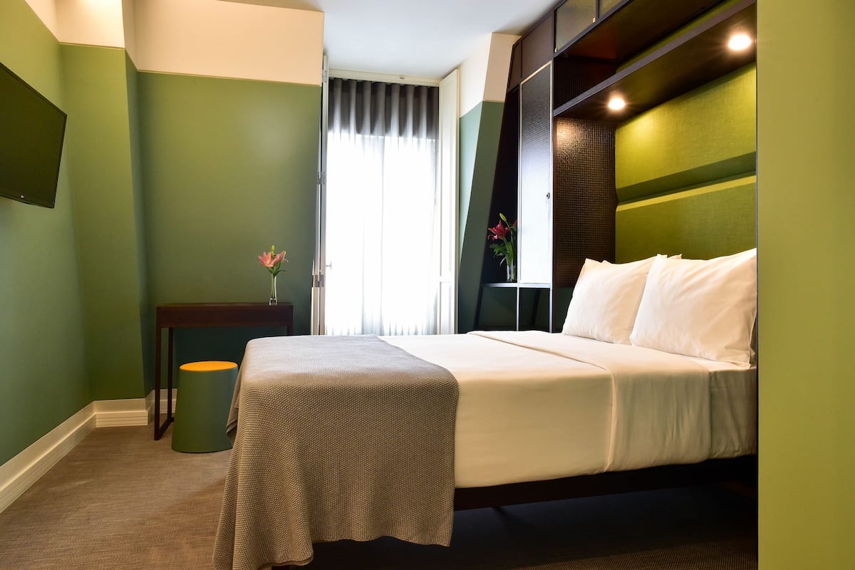 A hotel room with a bed, a nightstand, a small table, and a window with curtains.