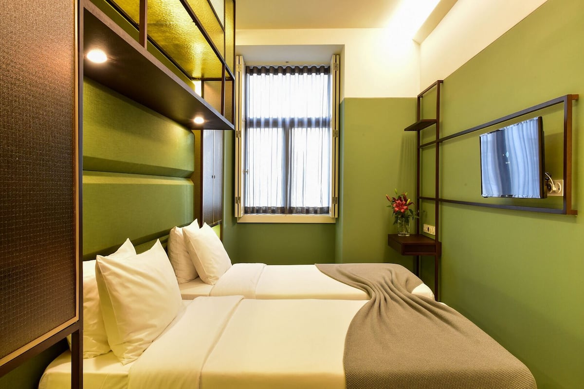 A hotel room with a bed, a window with curtains, a mirror, and a small table with a vase of flowers.