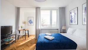 A hotel room with a large bed, a TV, a desk with a chair, and a window with curtains. - Riverside Comfort Studio (Warsaw)