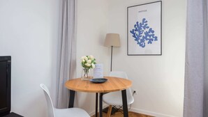 A small, neatly arranged room with a round wooden table, two white chairs, a vase with white flowers, a framed artwork on the wall, and a lamp. - Riverside Comfort Studio (Warsaw)