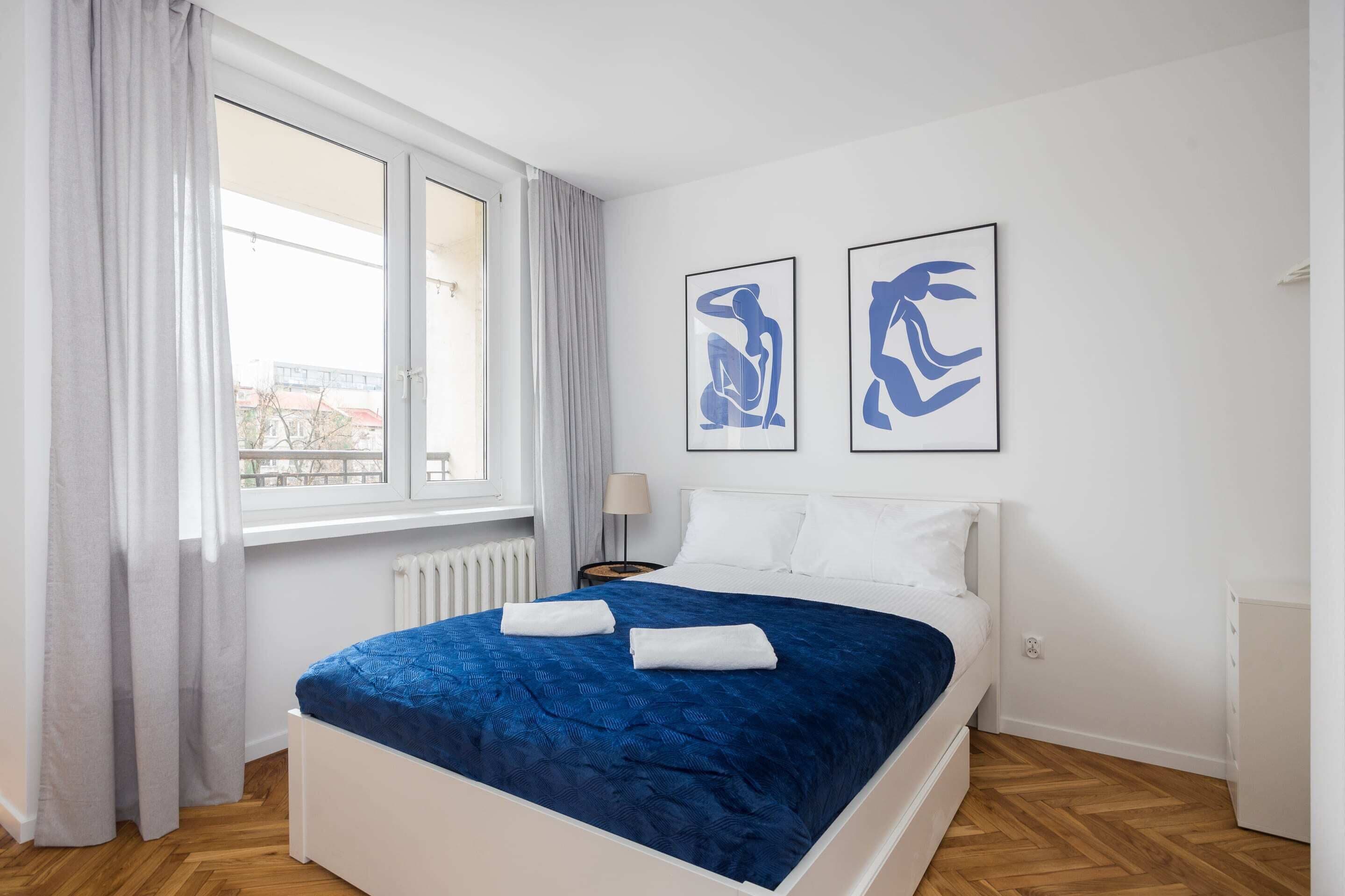 A bedroom with a bed, white curtains, a window, a radiator, a nightstand, a lamp, and two abstract art pieces on the wall.