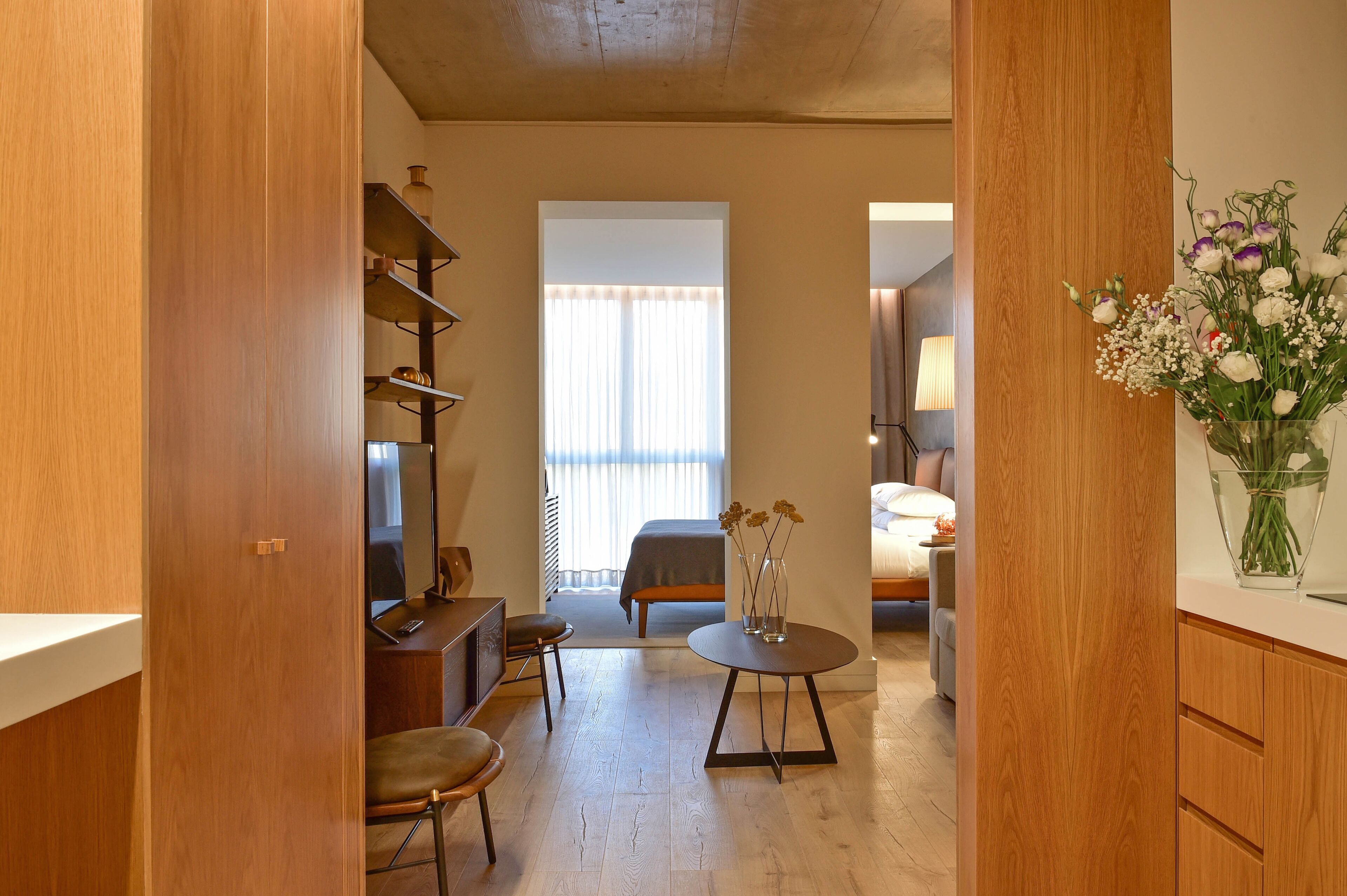 A modern room with a wooden floor, a bed with a gray headboard, a desk with a chair, and a small table with a vase of flowers.