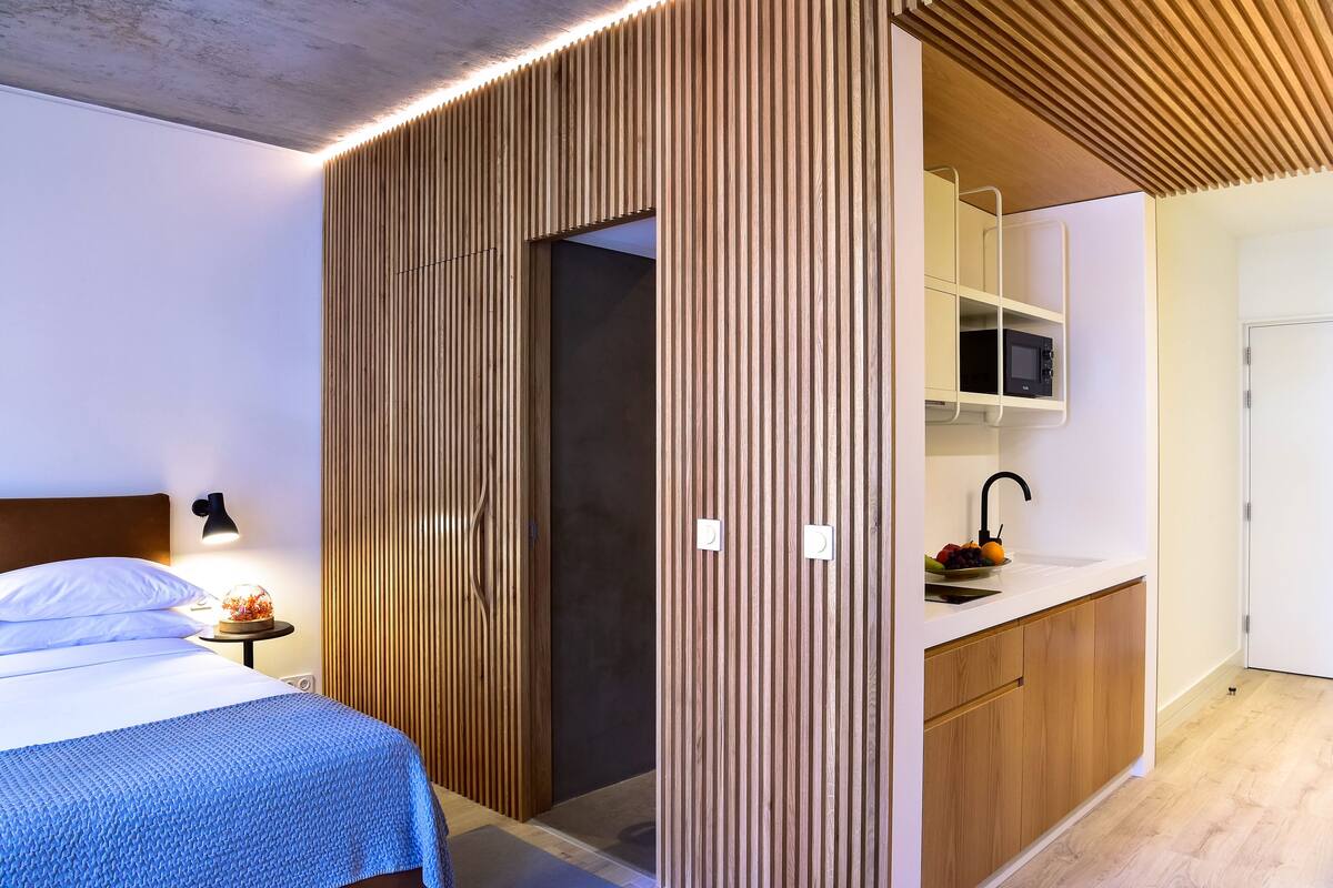 Ein modernes Hotelzimmer mit Bett, Nachttischlampe und einer Holzwand, die Schlafbereich und Küchenzeile trennt.