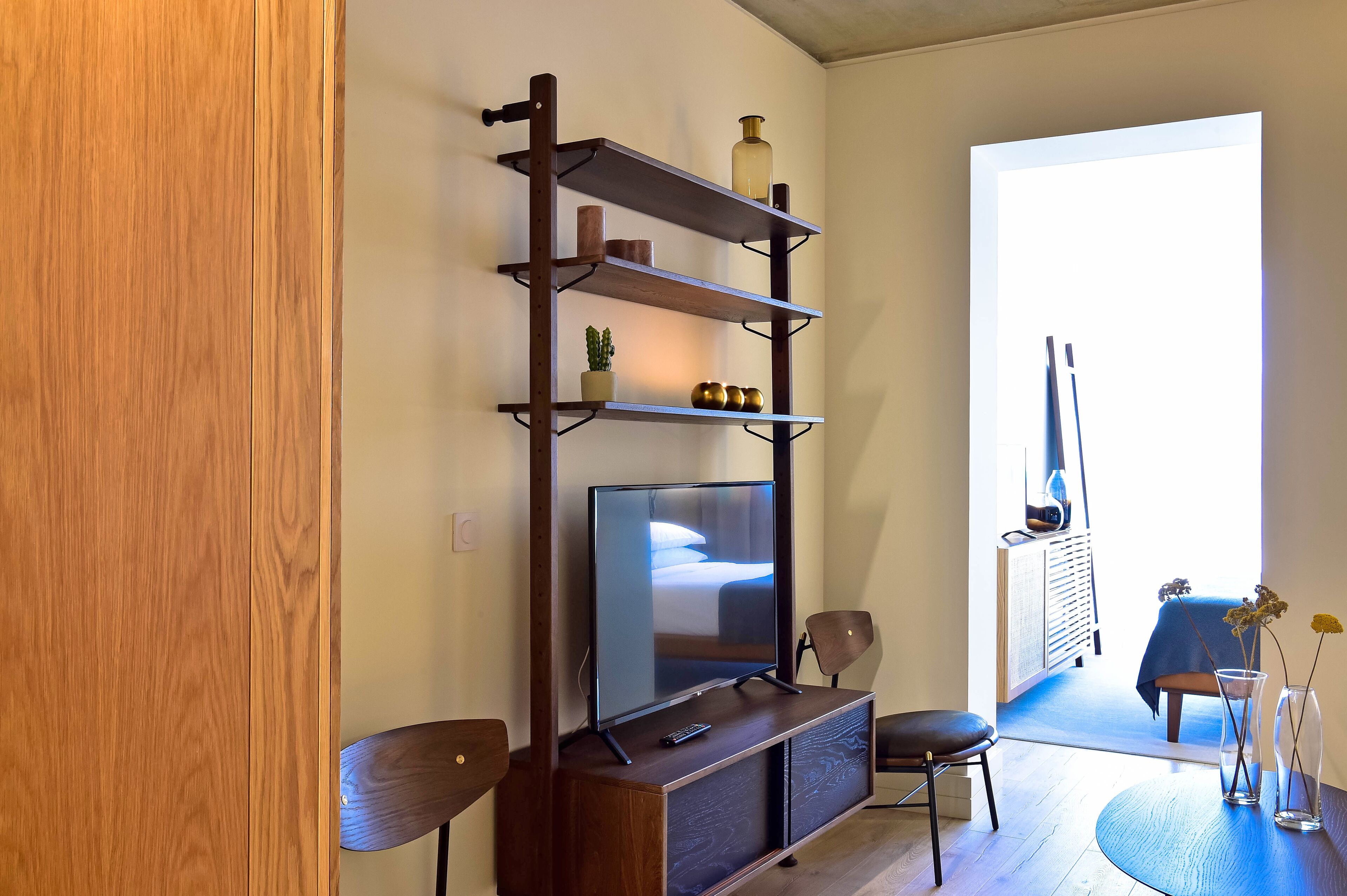 Ein modernes Wohnzimmer mit Fernseher auf einem Holzständer, einem kleinen Tisch mit einer Vase und einem Sessel.