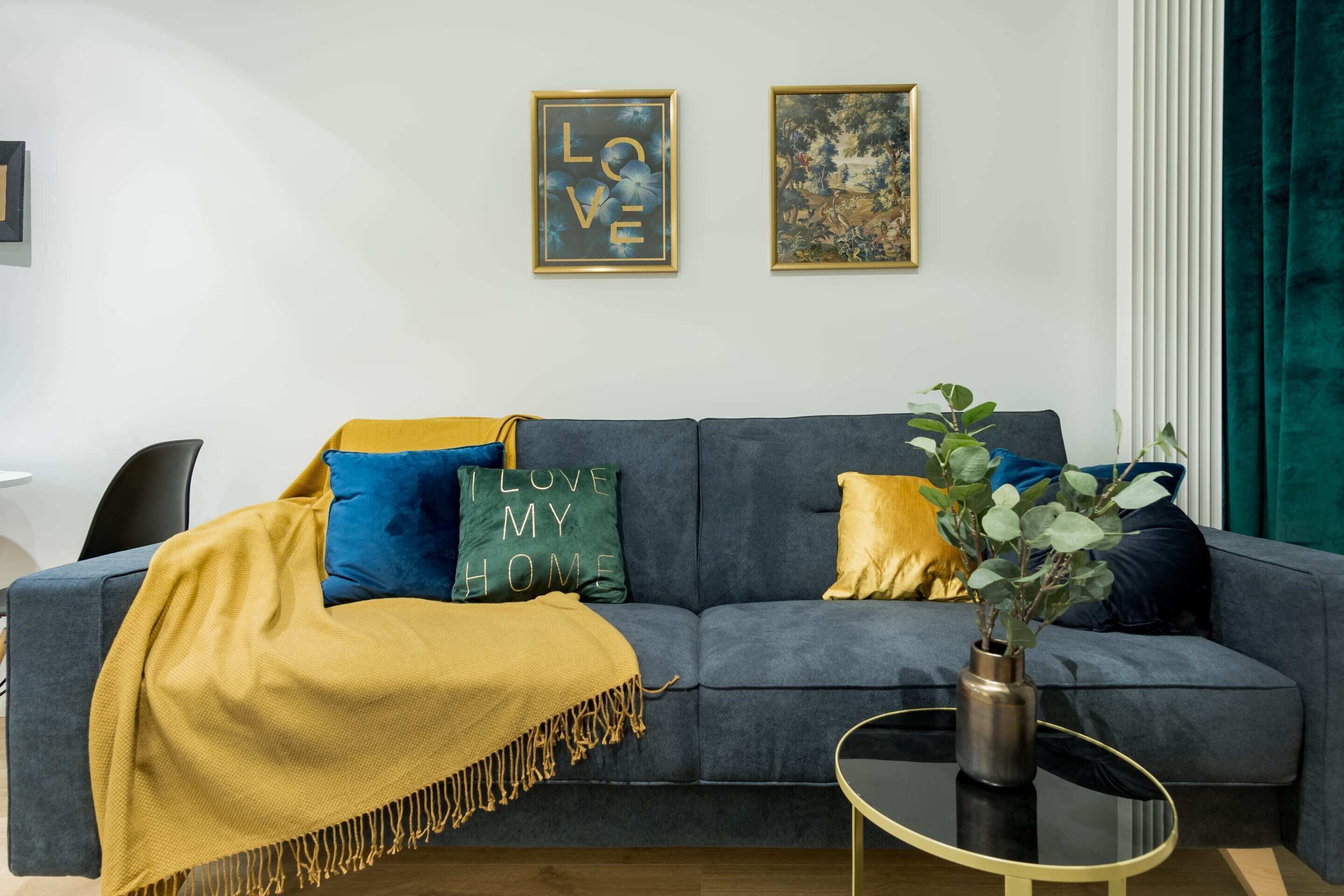 Un soggiorno moderno con un divano grigio, cuscini blu e dorati, una coperta gialla e due quadri appesi al muro.