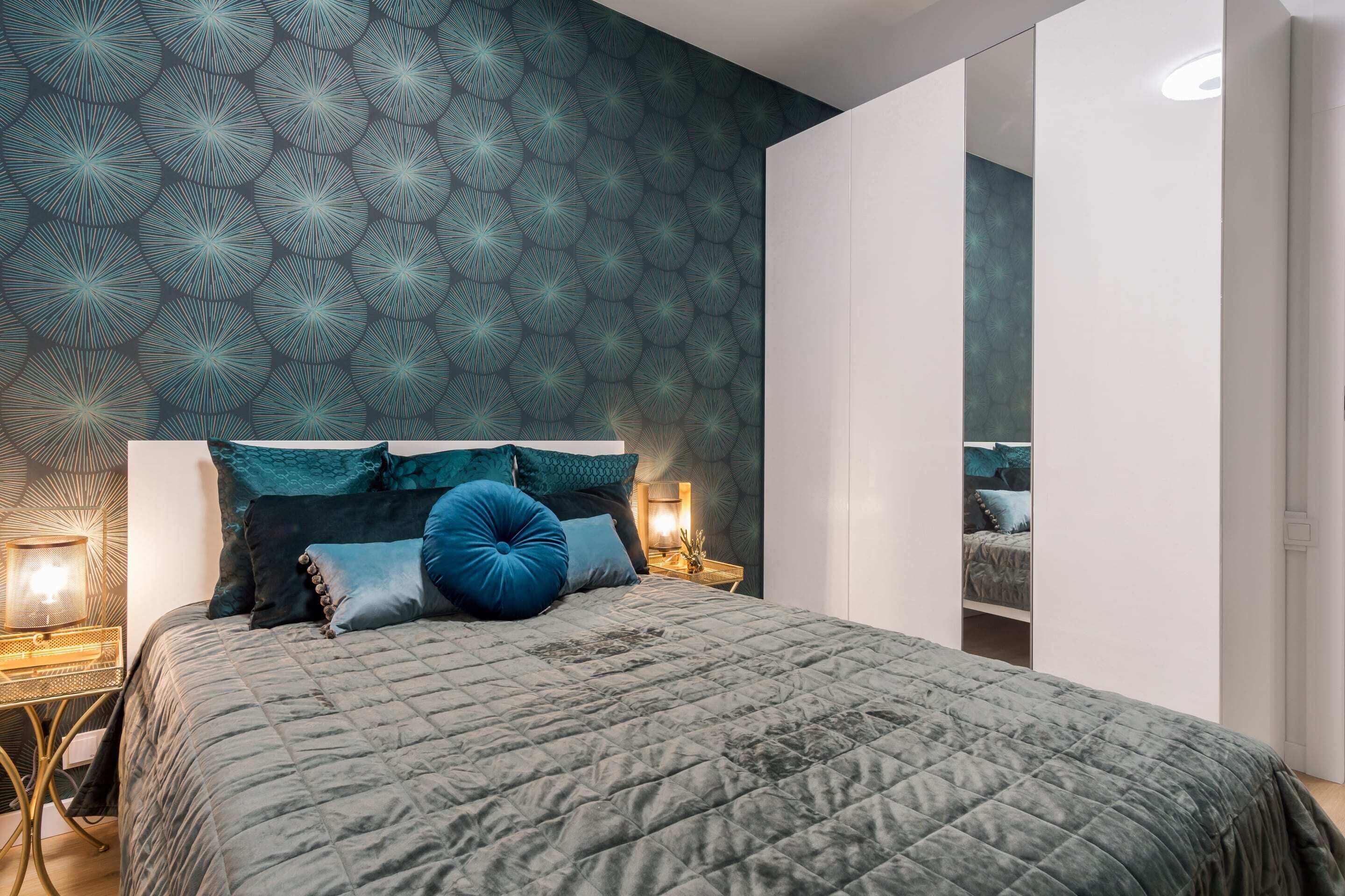 Un dormitorio moderno con una pared estampada, una cama grande con edredón gris acolchado y una mesita de noche con lámpara.