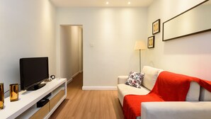A modern living room with a flat-screen TV on a white console, a red throw blanket on a white sofa, and a floor lamp. - Omar do Rio - SL109 (Rio de Janeiro)