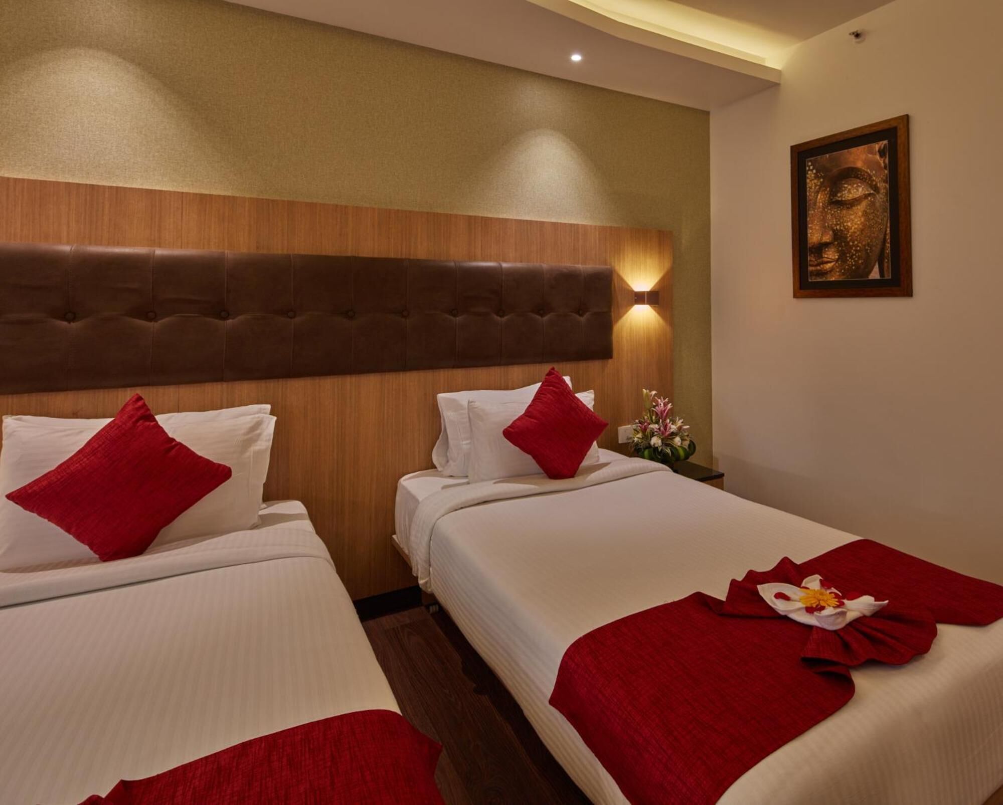 A hotel room with two beds, each with white linens and red pillows. There is a wooden headboard, a wall-mounted light, and a framed artwork above the beds.
