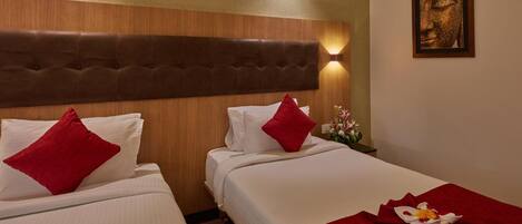 A hotel room with two beds, each with white linens and red pillows. There is a wooden headboard, a wall-mounted light, and a framed artwork above the beds.