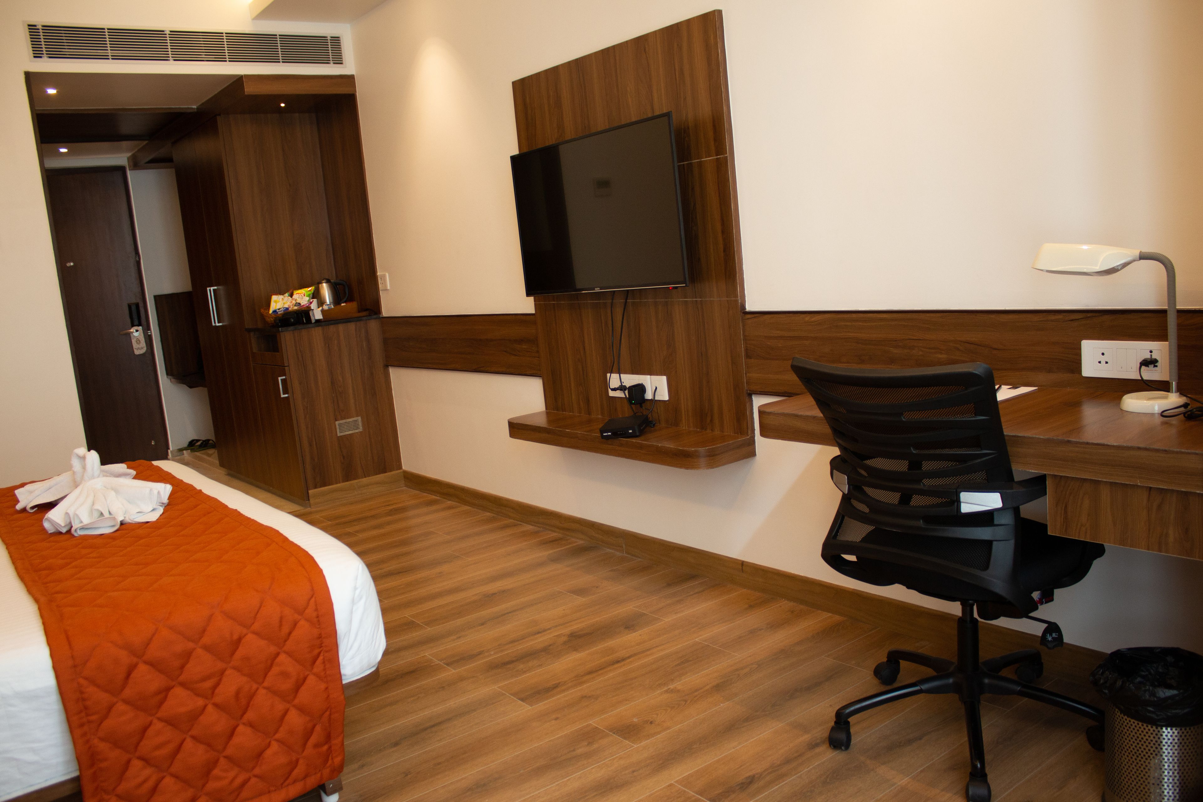 A hotel room with a bed, a desk, a chair, a TV mounted on the wall, and a trash bin.