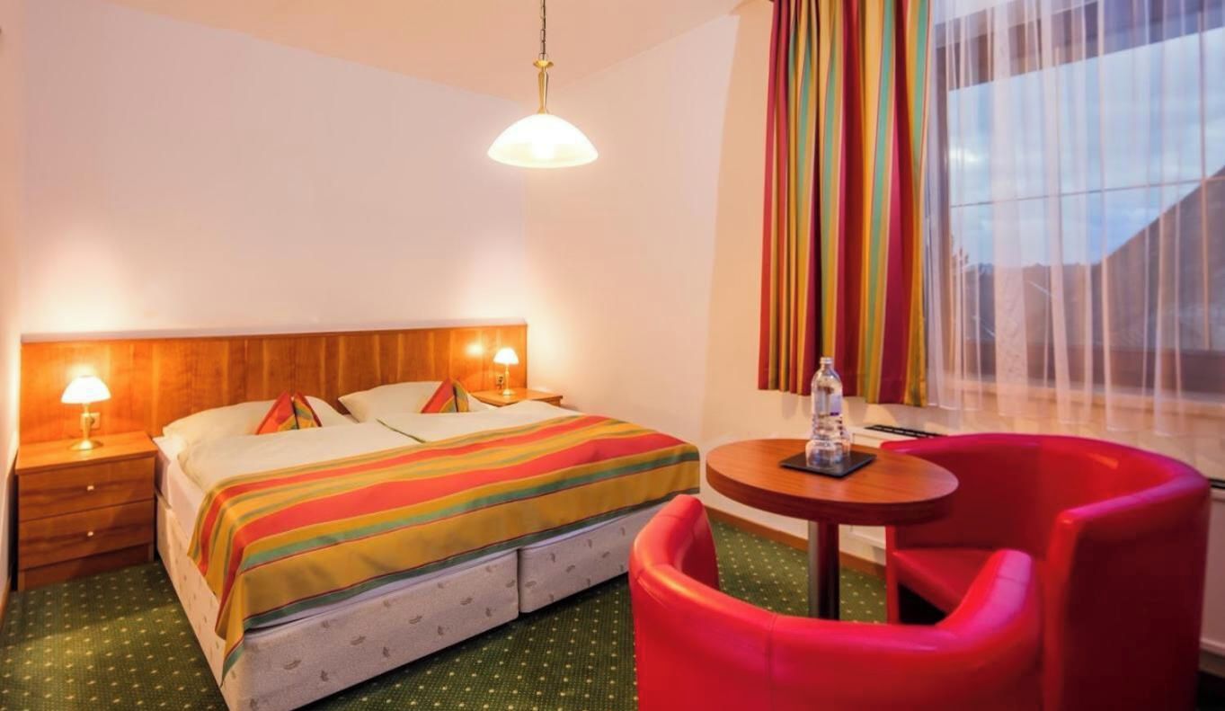A hotel room with a bed, two bedside lamps, a round table with a chair, and a window with curtains.