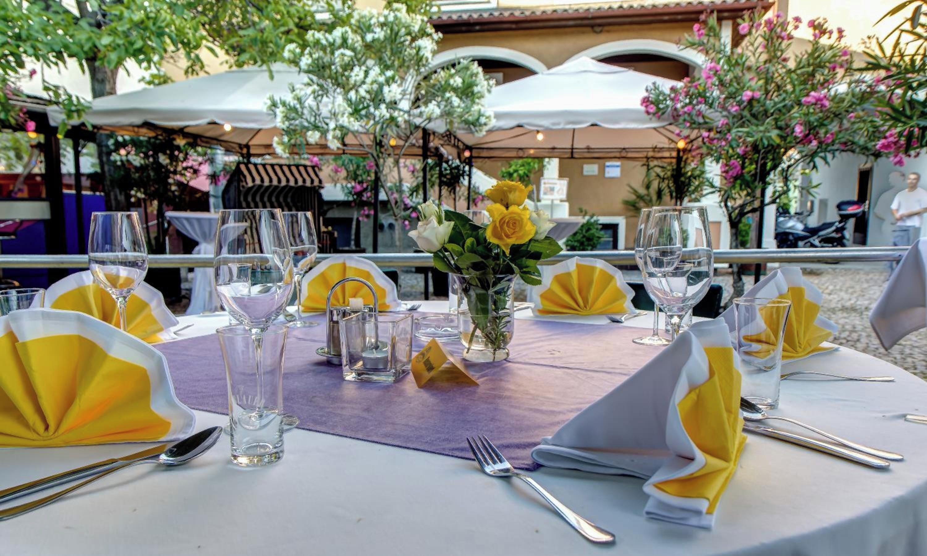 An outdoor dining area with tables set for two, featuring clear glassware, napkins, and a centerpiece of yellow roses.