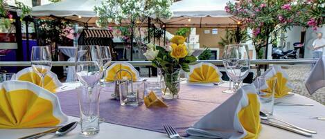 An outdoor dining area with tables set for two, featuring clear glassware, yellow napkins, and a vase with yellow roses.