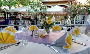 An outdoor dining area with tables set for two, featuring clear glassware, napkins, and a centerpiece of yellow roses.