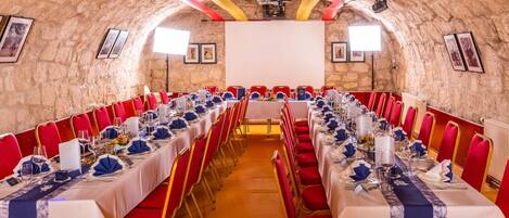 Un salón de conferencias con mesas largas, sillas rojas y una pantalla para proyector.