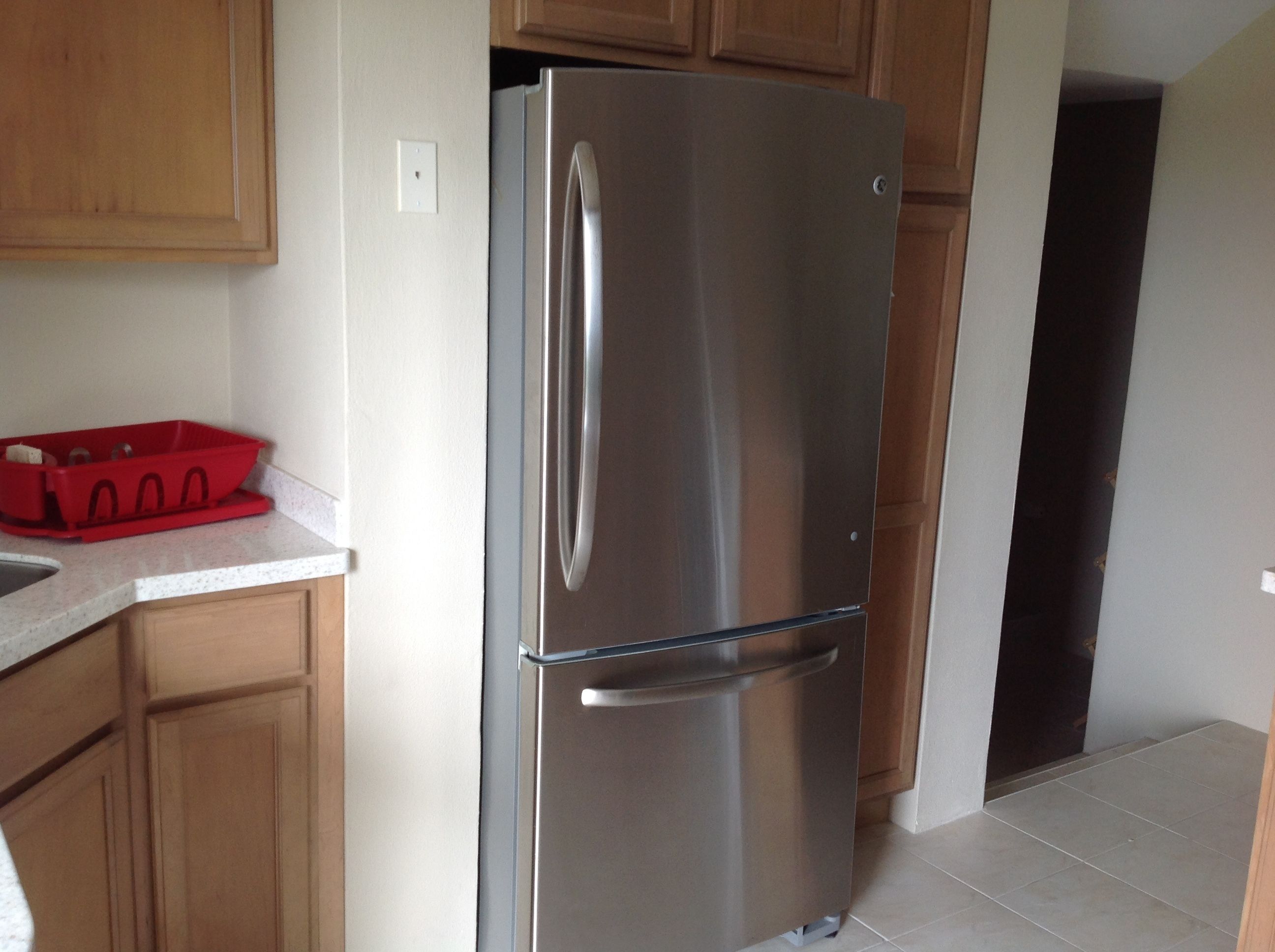 A stainless steel refrigerator in a kitchen with wooden cabinets and a red dish rack on the counter.
