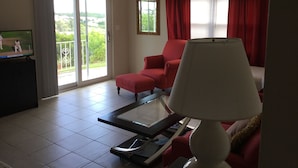 A living room with a television, a red armchair, a glass coffee table, a ceiling fan, and a view through a sliding glass door. - Central Apartment (Devonshire Parish)