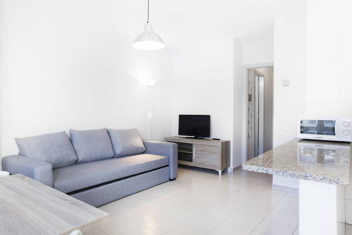Uma sala de estar moderna com um sofá cinzento, uma televisão de tela plana em um suporte de madeira e uma área de cozinha com micro-ondas.