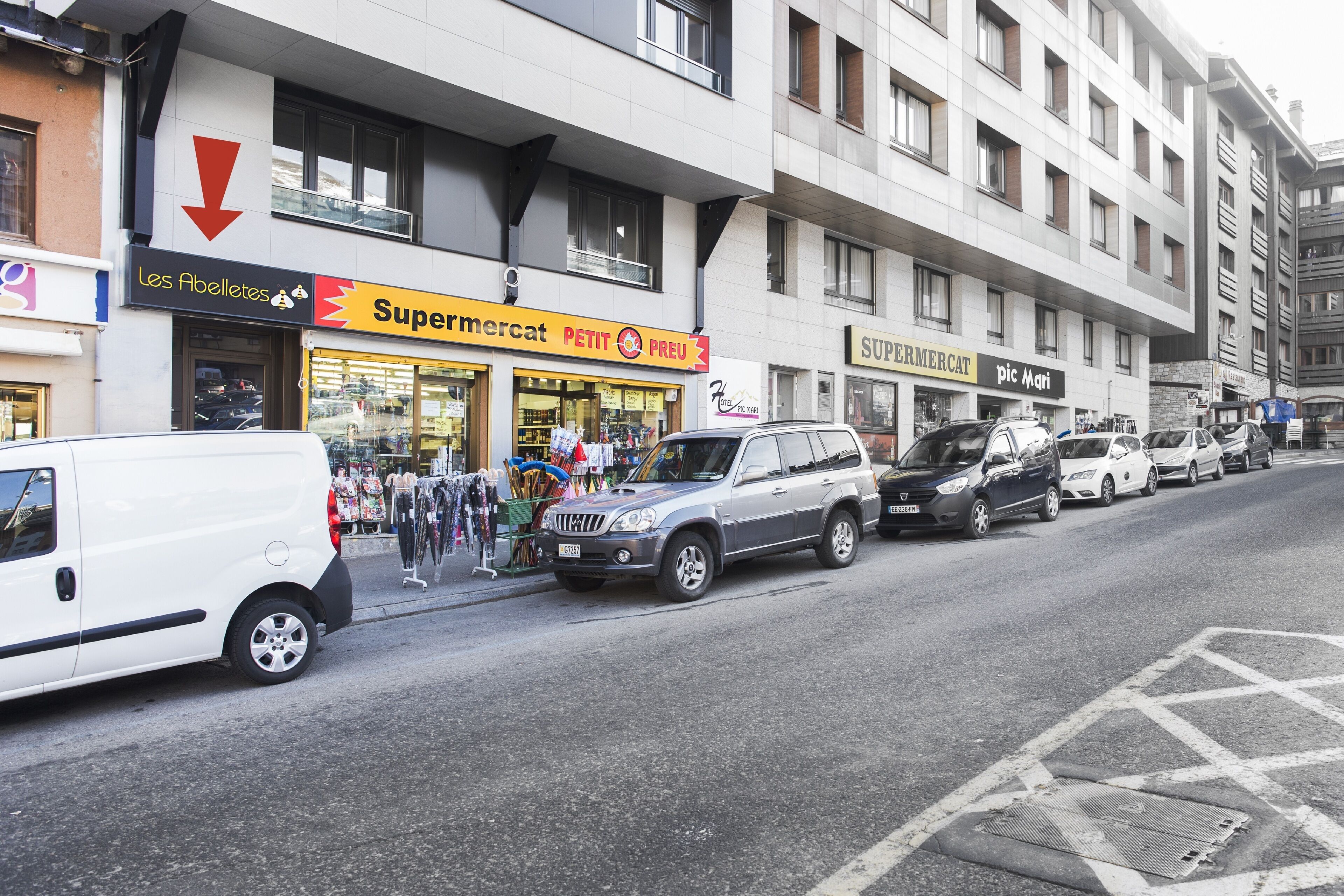 A street view with a row of parked cars and a building with a 'Supermercat' sign.