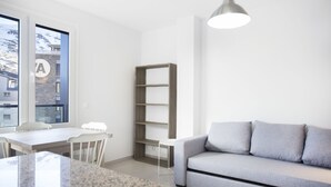 Een moderne woonkamer met een grijze bank, een houten boekenkast, een eettafel met stoelen en een groot raam met uitzicht op gebouwen.