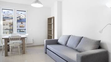 A modern living room with a grey sofa, a dining table with chairs, a wooden bookshelf, and a large window offering a view of buildings.